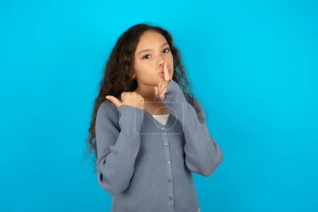 Téléchargez les photos : Adolescente portant un pull gris sur fond bleu demandant de se taire avec le doigt sur les lèvres pointant la main sur le côté. Silence et concept secret. - en image libre de droit