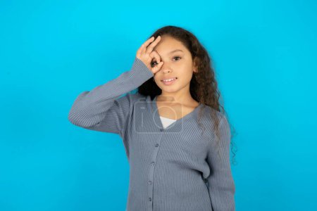 Photo for Teenager girl wearing grey sweater against blue background doing ok gesture with hand smiling, eye looking through fingers with happy face. - Royalty Free Image