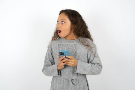 Foto de Adolescente usando suéter gris sosteniendo un teléfono inteligente y mirando hacia los lados en blanco copyspace. - Imagen libre de derechos