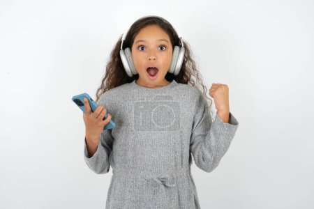 Foto de Chica adolescente positiva con suéter gris sostiene teléfono celular moderno conectado a los auriculares, cierra el puño de las buenas emociones, exclama con alegría, - Imagen libre de derechos