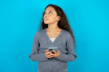 Téléchargez les photos : Adolescent fille portant pull gris tenant dans les mains téléphone mobile et regardant de côté avec look rêveur. - en image libre de droit