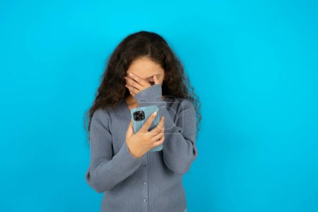 Photo for Teenager girl wearing grey sweater looking at smart phone feeling sad holding hand on face. - Royalty Free Image