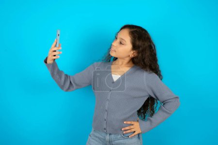 Foto de Adolescente chica usando gris suéter sonriendo y tomando un selfie listo para publicarlo en sus redes sociales. - Imagen libre de derechos