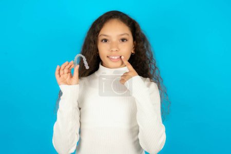Téléchargez les photos : Adolescente portant un pull blanc tenant un aligneur invisible et pointant des dents droites parfaites. Concept de soins dentaires et de confiance. - en image libre de droit