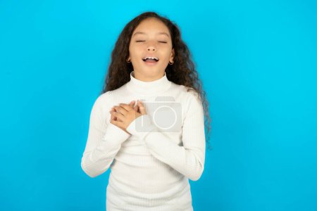 Teenager-Mädchen im weißen Pullover drückt Glück aus, lacht angenehm, hält die Hände am Herzen