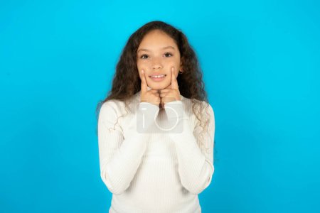 Foto de Chica adolescente feliz vistiendo suéter blanco con sonrisa dentada, mantiene los dedos índice cerca de la boca, dedos señalando y forzando sonrisa alegre - Imagen libre de derechos