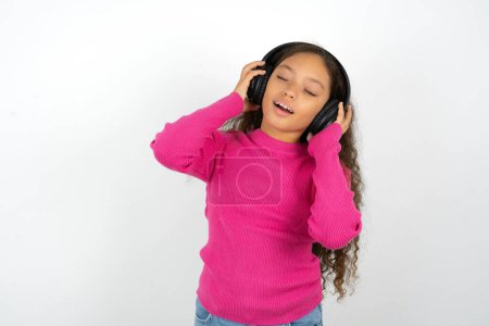 Téléchargez les photos : Adolescent fille portant pull rose sourit se sent généralement très heureux écoute piste de musique préférée via casque sans fil ferme les yeux. - en image libre de droit