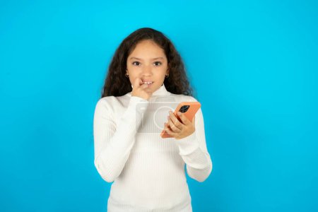 Photo for Afraid funny teenager girl wearing white sweater holding telephone and bitting nails - Royalty Free Image