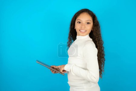 Téléchargez les photos : Photo de heureux joyeux intelligent jeune belle adolescente portant col roulé blanc sur fond bleu tenir tablette navigation sur Internet - en image libre de droit