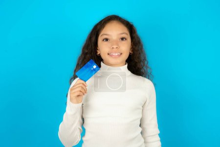 Photo for Photo of happy cheerful smiling positive young beautiful teen girl wearing white turtleneck over blue background recommend credit card - Royalty Free Image