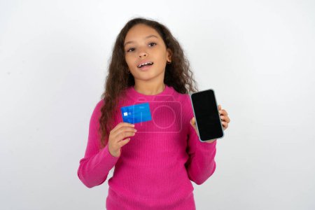 Foto de Adolescente con suéter rosa abrió cuenta bancaria, la celebración de teléfono inteligente y tarjeta de crédito, sonriendo, recomienda el uso de la aplicación de compras en línea - Imagen libre de derechos