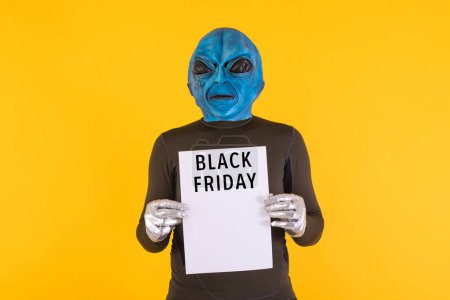 Foto de Alien con una cabeza azul sosteniendo un letrero blanco con las palabras 'Viernes Negro' en negro, sobre un fondo amarillo. Concepto de ofertas, descuentos, oportunidades, divertido, extraño y extraño. - Imagen libre de derechos