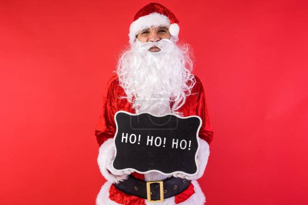 Téléchargez les photos : Homme habillé comme le Père Noël, tenant un signe noir qui dit : "Ho Ho Ho", sur un fond rouge. Noël, célébration, cadeaux, concept de consumérisme et de bonheur. - en image libre de droit