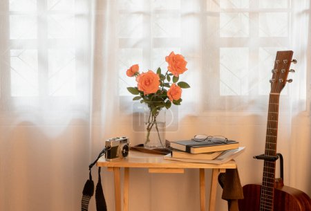 Foto de Rosas rosadas de coral en un jarrón, sobre la mesa hay libros y cuadernos, anteojos. y una cámara de cine decorada con una guitarra acústico-eléctrica marrón, fotografiada por la ventana de la habitación - Imagen libre de derechos