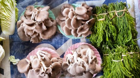 Photo for Pleurotus Pulmonarius mushrooms piled up in plates and acacia vegetables are sold on tables at the home market. - Royalty Free Image