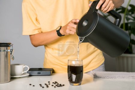 Foto de Man making use kettle pouring hot water making iced coffee or cold americano coffee into the cup with equipment, tool digital scale on the wooden bar at kitchen home - Imagen libre de derechos