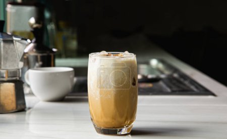 Téléchargez les photos : A glass of iced coffee with milk, Asian caffeinated beverages on a marble bar, and a Moka Pot coffee pot in the kitchen. - en image libre de droit