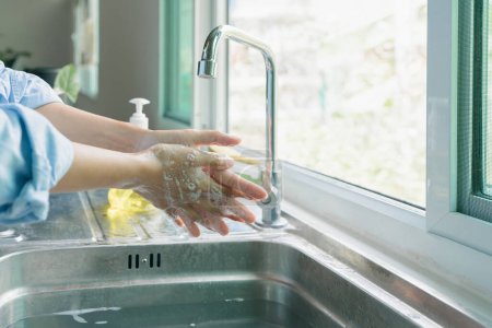 Foto de Mujer asiática de primer plano bronceada a mano con camisa azul lavándose ambas manos. limpie su dedo con agua y jabón de espuma en el fregadero de la cocina antes de comer. concepto de seguridad germinal - Imagen libre de derechos