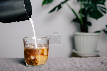 Téléchargez les photos : Close-up, black stainless steel pitcher pouring frothed milk over mug into black coffee with ice mug on gray bar with potted plants background. - en image libre de droit