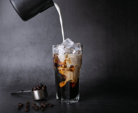 Foto de Pour milk make latte coffee into a transparent glass with ice. and roasted coffee beans on a black background studio photo - Imagen libre de derechos