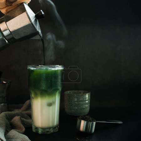 Photo for Poured Matcha green tea with Italian Moka pot pouring into a cup with iced of milk and powdered green tea on spoon on black background. copy space for your text - Royalty Free Image