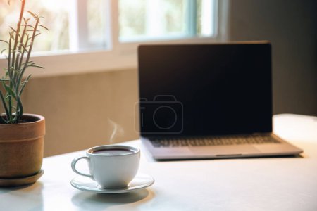 Téléchargez les photos : Tasse de café arôme sur la table avec ordinateur portable bureau à domicile le matin, boisson énergisante avant de commencer le concept de travail. gros plan - en image libre de droit