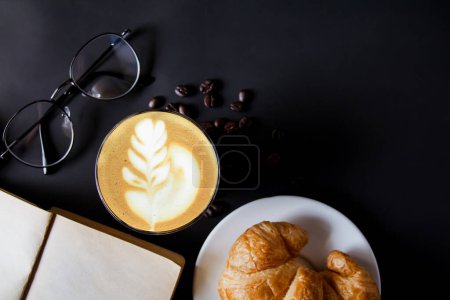 Foto de Taza de café caliente o café latte art Granos de café tostados y croissants con gafas de utilería, libro sobre fondo negro, fotografiado en el estudio. Vista superior - Imagen libre de derechos