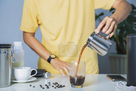Foto de Gente con camisa amarilla vertiendo café americano en un vaso transparente con hielo. y frijoles de café tostados y equipo herramienta de elaboración enblanco de la mesa. hacer café en casa - Imagen libre de derechos