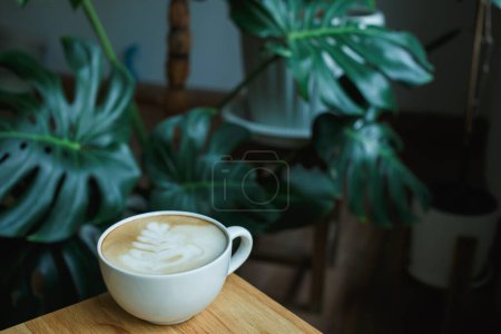 Foto de Enfoque selectivo arte latte casero de café caliente en mesa de madera con árbol de monstera, decoración de maceta de plantas en casa en el café de la mañana - Imagen libre de derechos