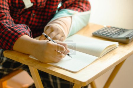 Téléchargez les photos : Les mains d'un étudiant masculin écrivaient des devoirs de mathématiques dans un cahier avec la calculatrice sur la table et son bras droit a été mis dans une attelle. - en image libre de droit