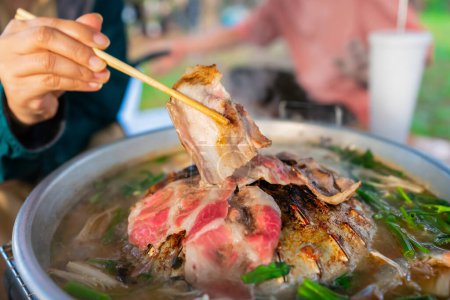 Korean BBQ or MOOKATA with sliced pork and vegetable soup, People hand using chopsticks grilled dinner the family. picnic outdoor. Close up	