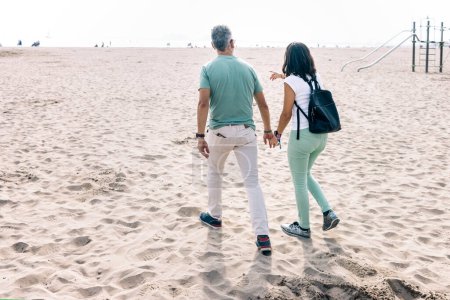 Adult tourist couple touring vacation beach. Man holding woman hand while walking along beach on sunny day. Husband and wife strolling along coast side area of their city.