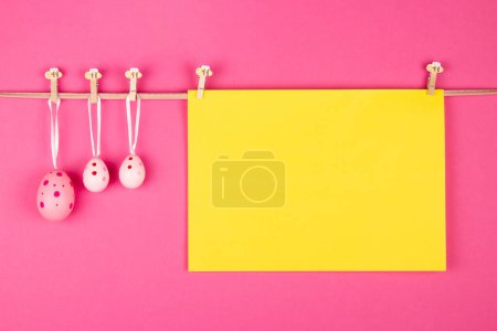 Téléchargez les photos : Feuille de papier jaune vierge et trois œufs de Pâques roses accrochés à la corde sur fond rose. Concept de carte de vœux de Pâques avec espace libre pour écrire - en image libre de droit
