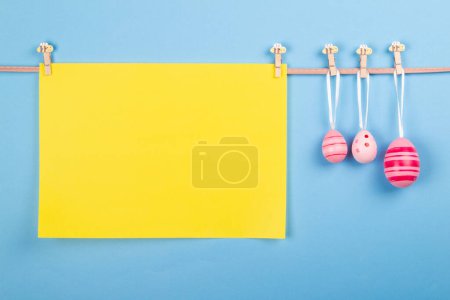 Téléchargez les photos : Feuille de papier jaune vif vierge et trois œufs de Pâques roses accrochés à la corde sur fond bleu clair. Thème carte de vœux de Pâques avec espace pour la personnalisation - en image libre de droit