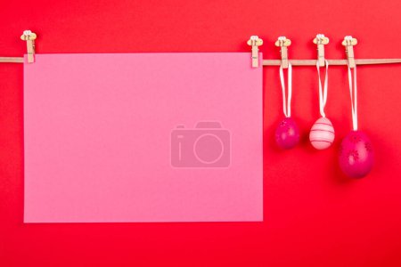 Téléchargez les photos : Feuille de papier de couleur rose vierge et trois œufs de Pâques roses accrochés à la corde sur fond rouge. Modèle de carte de Pâques avec zone vierge pour le texte - en image libre de droit