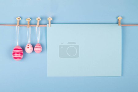 Téléchargez les photos : Feuille de papier bleu pastel vierge et trois œufs de Pâques roses accrochés à la corde sur fond bleu clair. Carte de Pâques avec une zone désignée pour écrire - en image libre de droit