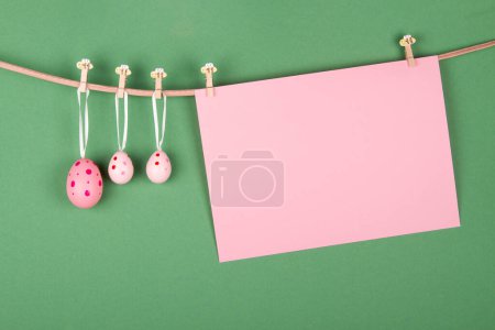 Téléchargez les photos : Feuille de papier rose vierge et trois œufs de Pâques roses accrochés à la corde sur fond vert. Conception de carte de vœux de Pâques avec espace vide pour le texte - en image libre de droit