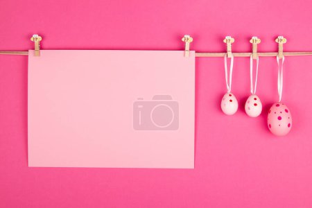 Téléchargez les photos : Feuille de papier rose pastel vierge et trois œufs de Pâques roses accrochés à la corde sur fond rose vif. Conception de carte de Pâques avec espace ouvert pour un message personnalisé - en image libre de droit