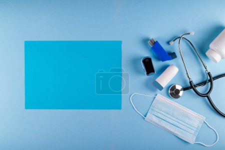Téléchargez les photos : Vue du dessus du masque médical, stéthoscope, oxymètre, bouteille de pilules et inhalateurs sur fond bleu clair avec feuille rectangulaire bleue vibrante de papier pour le placement de texte. Symptômes respiratoires asthmatiques Concept - en image libre de droit