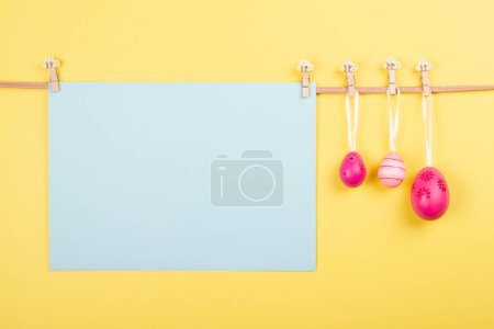 Téléchargez les photos : Feuille de papier bleu clair vierge et trois œufs de Pâques roses accrochés à la corde sur fond jaune pastel. Carte de Pâques avec espace ouvert pour vos mots ou votre image - en image libre de droit