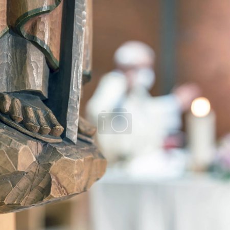 Heilige Holzstatue. Geschnitzte Holzfüße, Detail einer Holzstatue eines Heiligen. Im unscharfen Hintergrund feiert ein Priester die Messe.