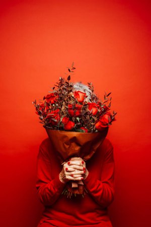 Senior woman wearing red clothes, hidding her face with a red roses bouquet, over a red background
