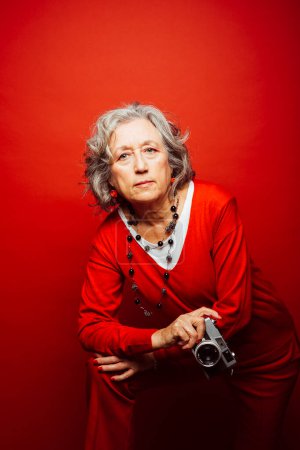 Photo for Senior woman wearing red clothes, taking pictures with an old film camera, over a red background - Royalty Free Image