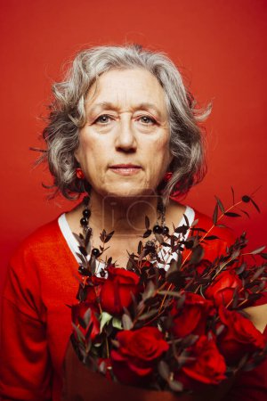 Photo for Senior woman wearing red clothes, holding a red roses bouquet, over a red background - Royalty Free Image