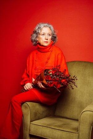 Photo for Senior woman wearing red clothes and sitting on a green armchair with a bouquet of red roses, over a red background - Royalty Free Image