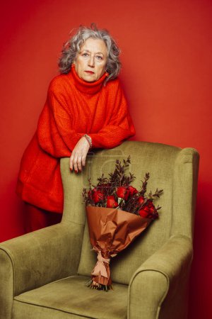 Photo for Senior woman wearing red clothes, standing next to a green armchair over a red background - Royalty Free Image