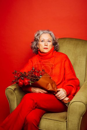 Photo for Senior woman wearing red clothes and sitting on a green armchair with a bouquet of red roses, over a red background - Royalty Free Image