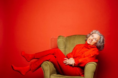 Photo for Senior woman wearing red clothes and sitting on a green armchair over a red background - Royalty Free Image