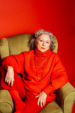 Photo for Senior woman wearing red clothes and sitting on a green armchair over a red background - Royalty Free Image