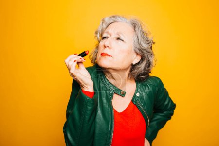 Retrato de una mujer mayor que aplica lápiz labial rojo sobre un fondo amarillo